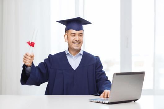 Graduate in computer technology holding diploma, celebrating success at home, with laptop on table.