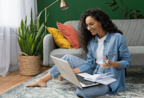 Laptop computer and notebook on a woman's lap, studying or working at home in a cozy living room with plants and colorful cushions, representing online education and career training at MyComputerCareer.