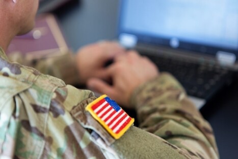 Soldier in uniform working on a computer with American flag patch on sleeve, representing cybersecurity or tech training for military personnel.